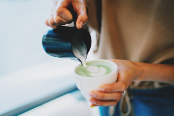 Barista making patterns while  making tasty and healthy matcha latte tea. Female's hand pouring hot whipped milk into the cup with freshly made natural matcha tea in a modern asian coffee shop.
