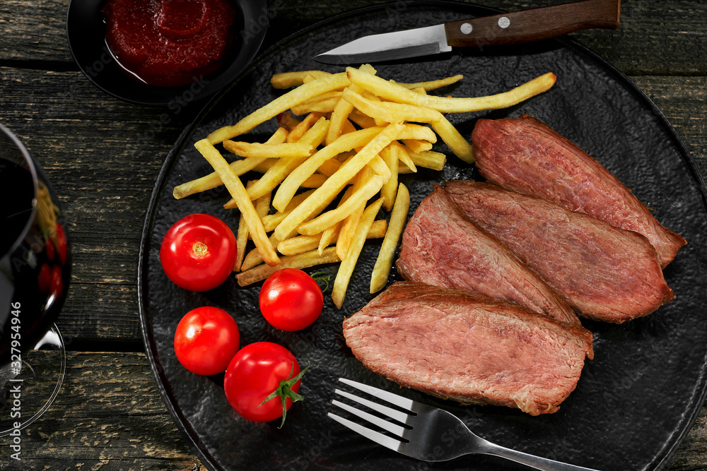 Wall mural Delicious medium rare steak with French fries and fresh cherry tomatoes on a black plate. Classic meat meal with red wine glass on a table. Top view, directly above shot.