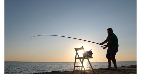 A fisherman reeling a fishing line on a reel, holding a fishing rod and straightening the fishing line at the sunrise, camera moving upwards from thermal box to a fisherman.