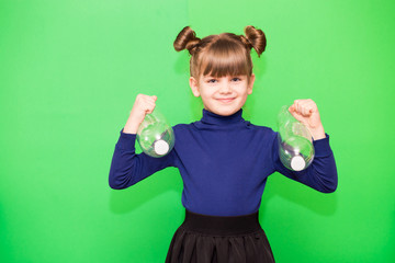 Positive little girl with funny pigtails shows her power biceps with arms raised victory holding plastic bottles isolated on green background
