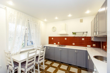 Modern stylish large kitchen in the cottage. Kitchen in red and steel colors. The doors are steel-colored, and the tiles on the wall are red. The floor tiles are beige. White table with four chairs.