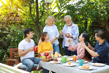 Happy multiethnic family standing and sitting at a breakfast table in backyard outdoor on sunny day with smiling face. Happy caucasian and asian family sitting and having conversation with good time.