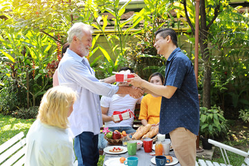 Obraz premium Happy multiethnic family giving surprise gift to caucasian grandfather on his happy birthday with happy smiling face in backyard outdoor on sunny day. Elder men shaking hands outdoor with happy face.