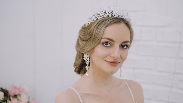 Close up tilt shot of young blonde bride with wedding hairstyle in tiara earrings wedding gown with bare shoulders posing in trendy wedding salon playful and seductively looking aside smiling playing