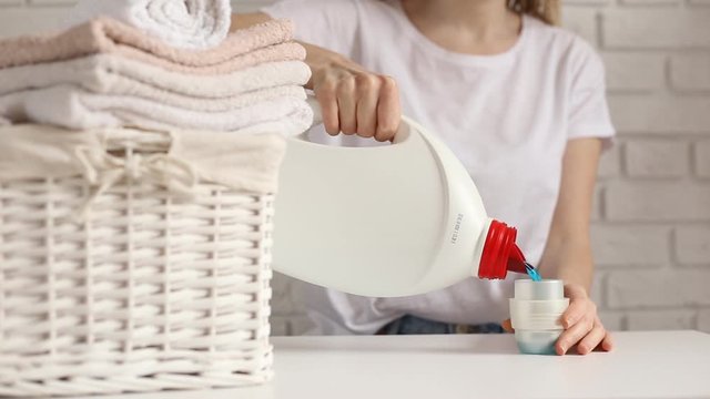 Close up of female hands pouring liquid laundry detergent into cap on white rustic table with towels in wicker basket