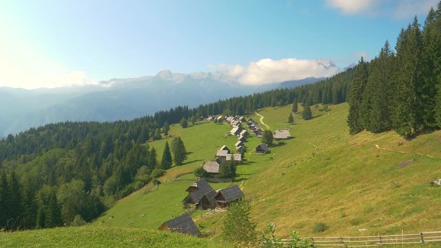 AERIAL: Scenic view of a small village of cottages with a view of the Julian Alps. Picturesque forest and untouched forest surrounds a row of wooden huts surrounding a hiking trail in Slovenia.