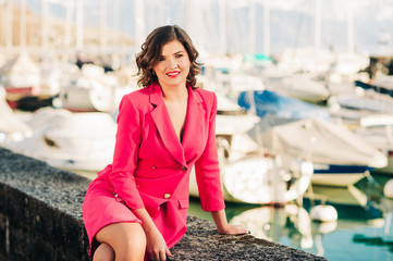 Outdoor fashion portrait of young stylish woman, resting by the lake, wearing pink jacket dress