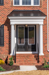 View of classic portico entrance covered w/ dark color metal roof, supported by two colonial white vinyl wrap columns w/ decorative caps and trim black baluster railing on a brick luxury American home