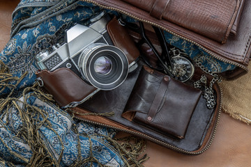 Top view of a vintage photo camera and a brown leather bag with scarf, glasses and pocket watch on sack cloth background.