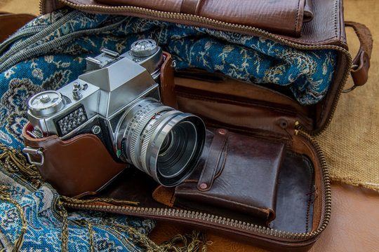 A vintage photo camera and a brown leather bag with scarf on sack cloth background.