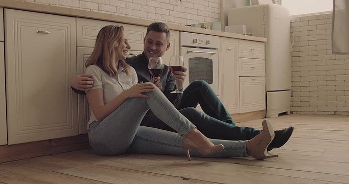Happy couple, Young man and woman sitting on floor in kitchen, drinking red wine and talking. Loving people spending time together at home.  4K slow motion video.