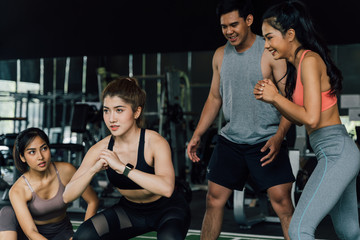 Fototapeta na wymiar Group of people cheering on their Asian female friend doing squats with a weight plate in fitness gym. Working out together as a teamwork. Encouragement and togetherness concept