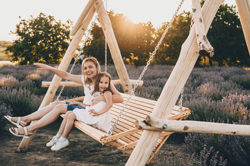 Lovely caucasian mother and her small daughter sitting in a swing with a lavender field on background - obrazy, fototapety, plakaty