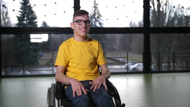 Close-up portrait of happy boy with cerebral palsy sitting in wheelchair in the middle of sport hall. Positive disabled teenager in eyeglasses and bright yellow T-shirt smiling at camera