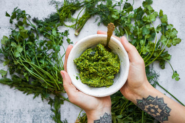 Green basil pesto paste dip sauce  in bowl with seeds, nuts and green herbs. Hands hold pesto. - obrazy, fototapety, plakaty