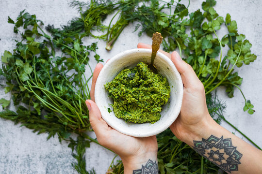 Green Basil Pesto Paste Dip Sauce  In Bowl With Seeds, Nuts And Green Herbs. Hands Hold Pesto.