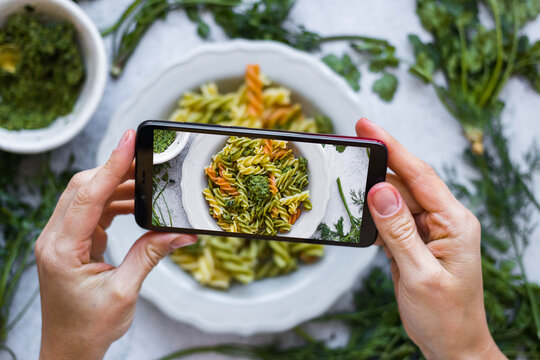 Make picture of food with phone. Hands hold smartphone. Photography of Italian pasta, macaroni with pesto and vegetables. Vegan meal. Social media and blogging content style. 