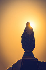 Silhouettes of the blessed Virgin Mary statue figure in a warm tone - sunset scene. Catholic praying for our lady - The Virgin Mary. - obrazy, fototapety, plakaty