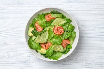 Salmon salad with cucumber, spinach, lollo, lettuce on dish. Healthy seafood, salmon fish, creative diet concept. Tasty salmon and mixed greens vegetarian salad on white wood, top view