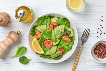 Salmon salad with cucumber, spinach, lollo, lettuce on dish. Healthy seafood, salmon fish, creative diet concept. Tasty salmon and mixed greens vegetarian salad on white wood, top view, flat lay