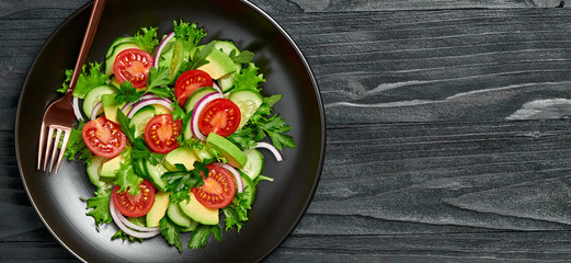 Fresh salad with cherry tomatoes, avocado,cucumeber and lettuce on dish. Healthy vegetarian meal concept. Tasty mixed leaves, tomatoes and mix vegetable salad on black wood, top view.