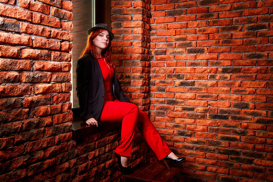 Beautiful young woman with redhair and black hat being near red brick wall inside