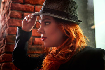 Beautiful young woman with redhair and black hat being near red brick wall inside