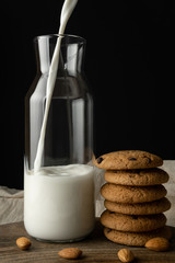 Oat cookies with chocolate, almond. Milk is poured into a transparent bottle on cutting board, over black background