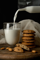 Oat cookies with pieces of chocolate, almond. Milk is poured into a glass with double walls from glass bottle. One cookie is broken. On cutting board