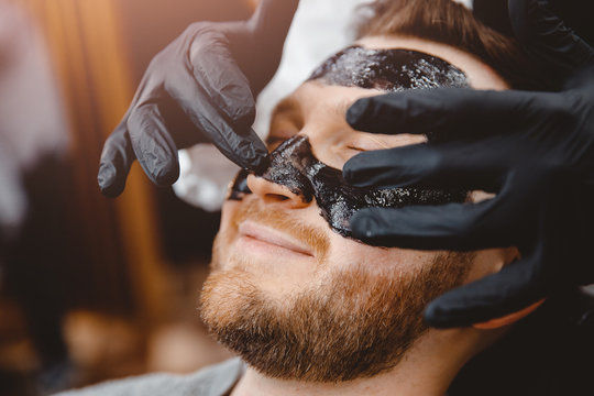 Barber applies black charcoal mask to man face to clean pore skin and remove acne from nose, spa salon