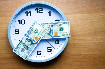 Analog clock and dollars on a wooden background. Banknotes are lying on the clock in the form of arrows. 100 dollars indicate hours and minutes. View from above. On the clock  time 20:15, 08:15