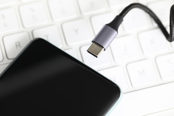 Close-up view of modern mobile phone and charger plug. Smartphone with black screen laying on white keyboard of laptop. Technology and usb battery concept