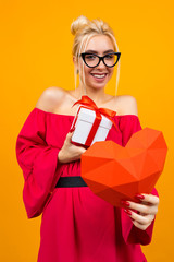attractive european girl in red dress holds gift box and red paper heart on orange studio background