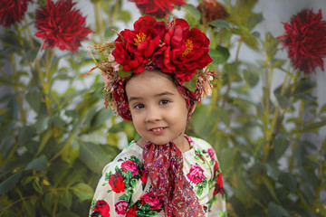 A little girl with a red floral crown sits under a flowering bush with red big flowers