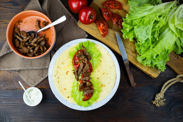 Burrito with slices of fried beef, tomato, lettuce, cheese tortilla. Final step of cooking lay the ingredients. Mexican national cuisine. Top view on food on the background of a wooden table.