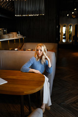 Business portrait of an attractive beautiful blonde woman in a blue jacket and white trousers in a cafe restaurant, a girl working , drinking coffee, talking on the phone, freelancing