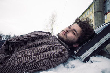 A bearded man in a coat lies on a car with a suitcase..
