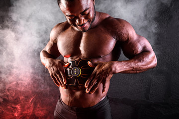 Bodybuilder male African uses electronic belt muscle stimulator trainer abdominal muscles on a black background with smoke - obrazy, fototapety, plakaty