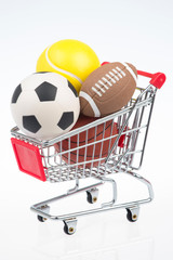 Shopping cart on white background filled with sporting goods including soccer ball, American football, tennis ball, and basketball