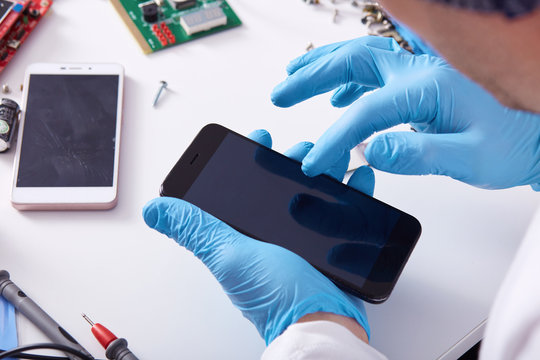 Close up faceless portrait of person's hand in blue rubber gloves fixing damaged screen of mobile phone, unknown person sitting at white tabble with different equipments. Modern technology concept.
