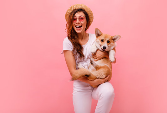 Cute brunette woman in white t shirt and jeans holding and embracing corgi puppy dog on plane pink background. Love to the animals, pets concept. cheerful woman holding Welsh Corgi dog. Happy together