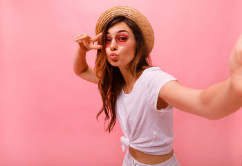 Portrait of a happy cheerful girl in summer hat showing peace gesture and making cute selfie...