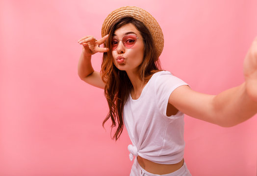 Portrait of a happy cheerful girl in summer hat showing peace gesture and making cute selfie isolated over pink background. Copy space