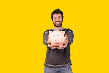 young handsome man with a piggy bank over yellow background, making funny gestures