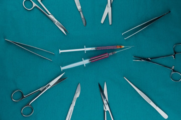 Top view of surgical instrument, liquid parenteral products in syringe with scalpel, tweezers, scissors on surgical green drape fabric.