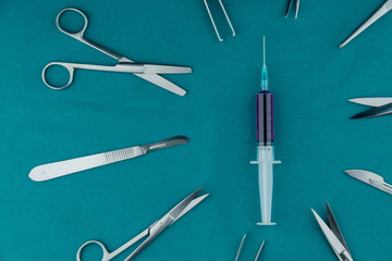 Top view of surgical instrument, liquid parenteral products in syringe with scalpel, tweezers, scissors on surgical green drape fabric.