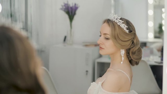 Close up shot of young blonde bride with wedding hairstyle in chic tiara earrings wedding dress with bare shoulders posing in front of makeup artist mirror in trendy wedding salon playful and