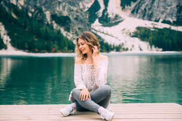 Fototapeta premium Beautiful girl long flowing hair sits on wooden pier, legs lotus position, hand near head smiles enjoy nature relax silence beauty of alpine Lake Braies. Backdrop white mountains green forest water