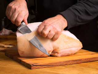 A male chef cuts a piece of pork carcass on a cutting board with a large knife.