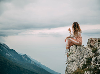 Red hair girl turned away in pink suit sits on edge mountain rock on backdrop sky clouds mountains foggy haze. Woman enjoy beauty fairytale nature silence relaxation. Top Peak Monte Baldo Alps Italy